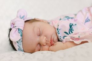 Sleeping baby wearing floral headband and pink outfit on white textured surface.
