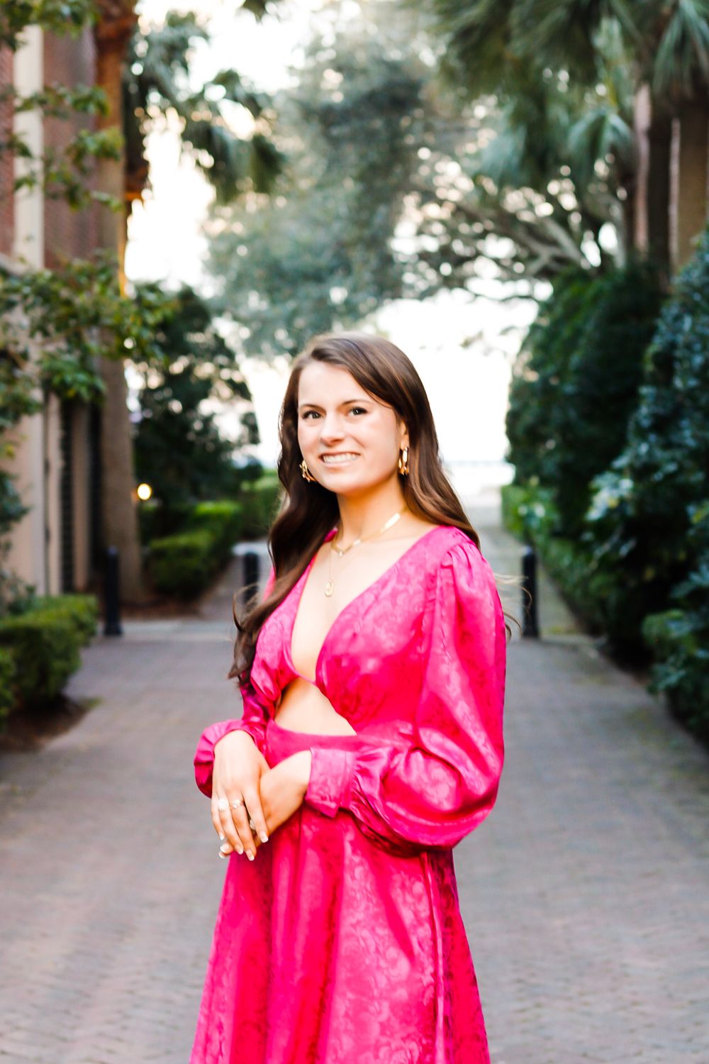 A smiling brunette woman in a vibrant pink satin dress stands on a tree-lined brick pathway.
