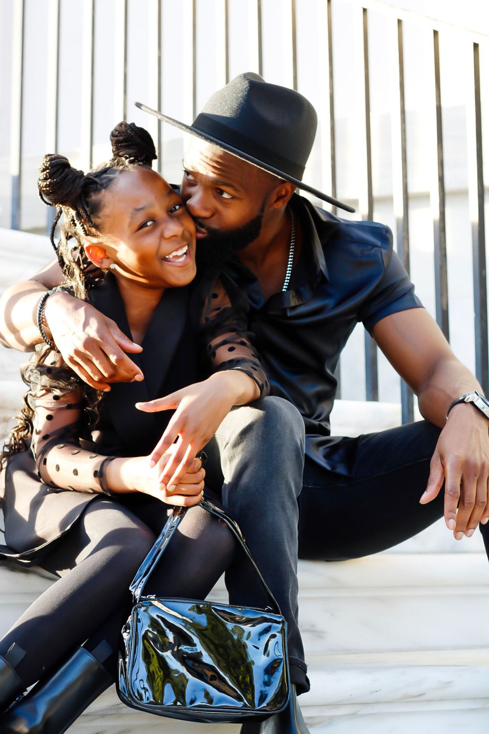 A man in a black hat kisses a smiling girl on the cheek while sitting together on steps.