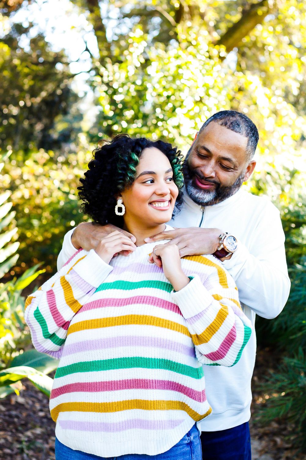 A couple embraces outdoors, woman in colorful striped sweater smiles as man in white hoodie wraps arms around her.