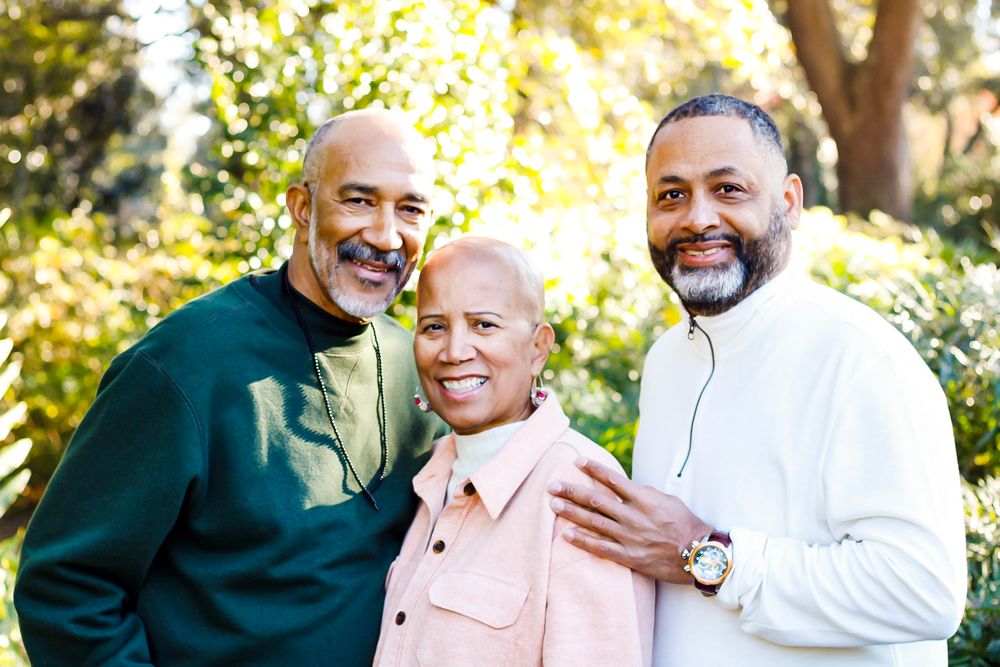 Three smiling Black adults pose together outdoors surrounded by lush green trees in a warm family portrait.