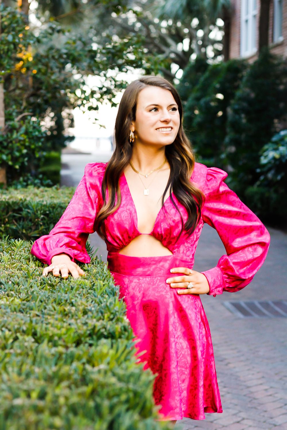 A brunette woman in a hot pink cutout dress poses gracefully among green hedges in an outdoor setting.
