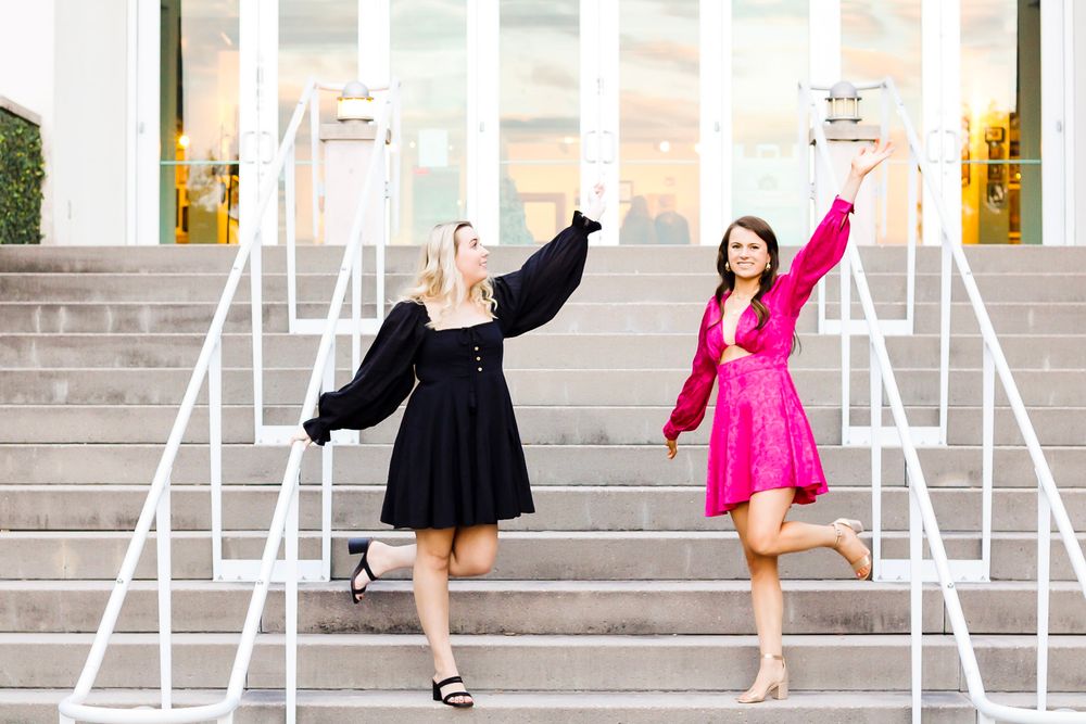 Two women in black and pink dresses celebrate on outdoor steps, raising books joyfully in the air.