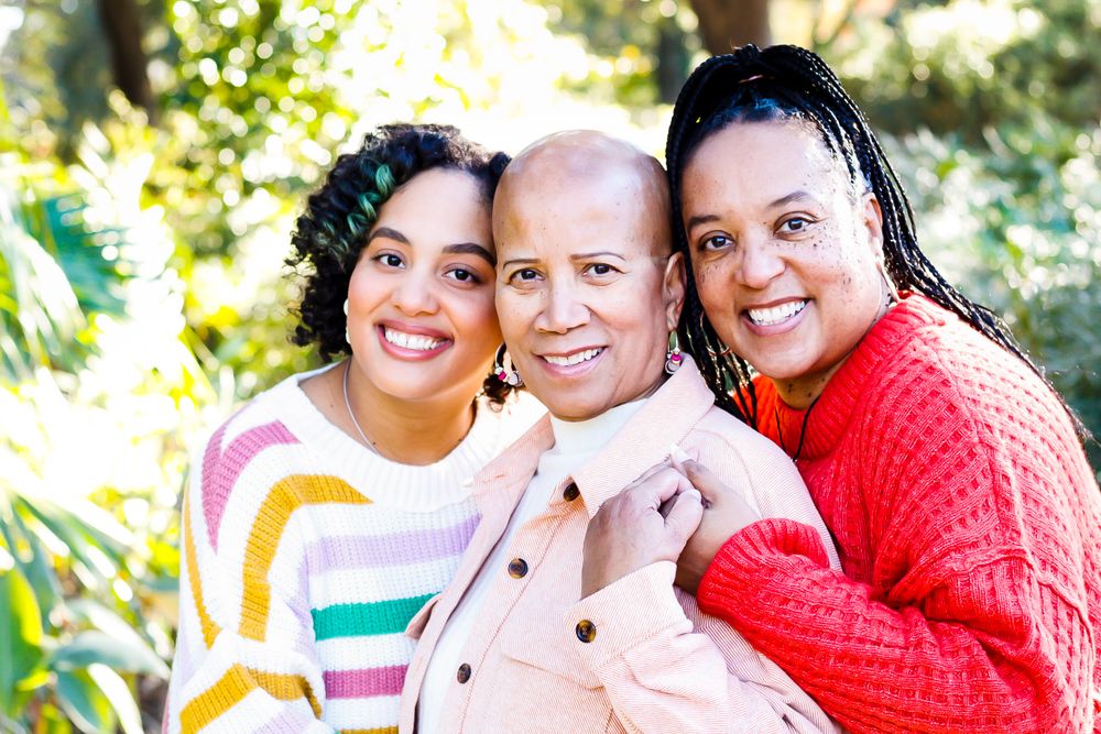Three Black women of different generations smiling and embracing outdoors among green foliage.