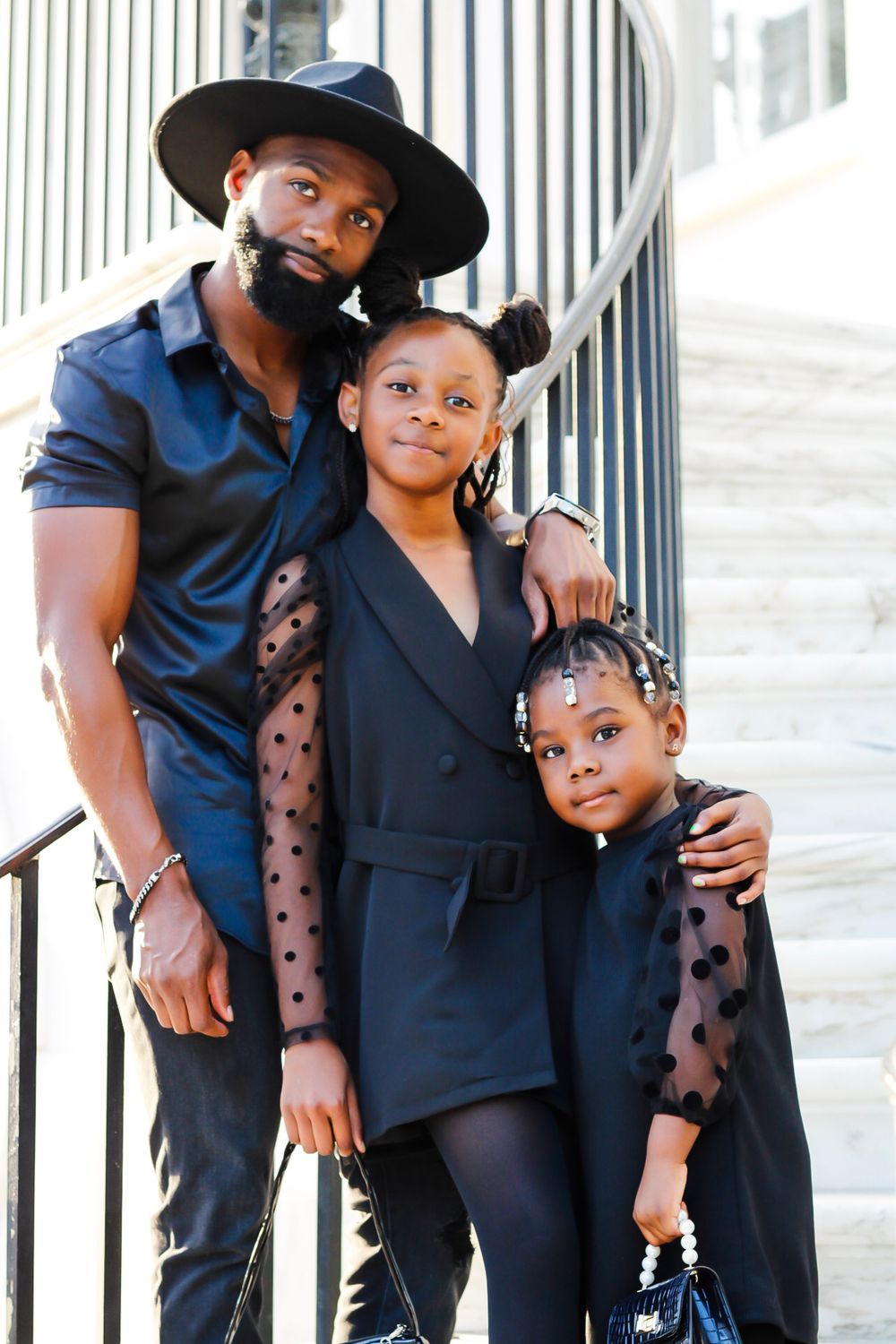 A father in all-black with a wide-brim hat poses with his two daughters on a staircase.