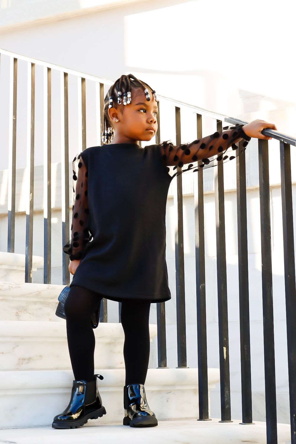 A young girl in a black dress with sheer polka-dot sleeves poses on white stairs holding a railing.