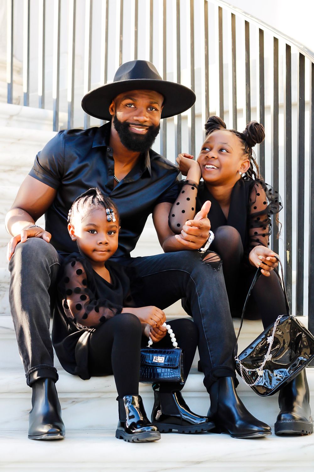 A father in a black hat sits on steps with his two daughters, all dressed in coordinating black outfits.