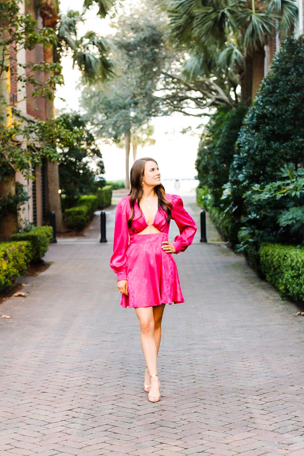 A brunette woman in a hot pink long-sleeve mini dress walks confidently down a tree-lined brick pathway.