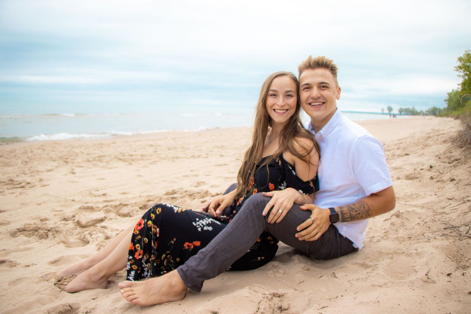 A couple sits together on a sandy beach with the ocean behind them.
