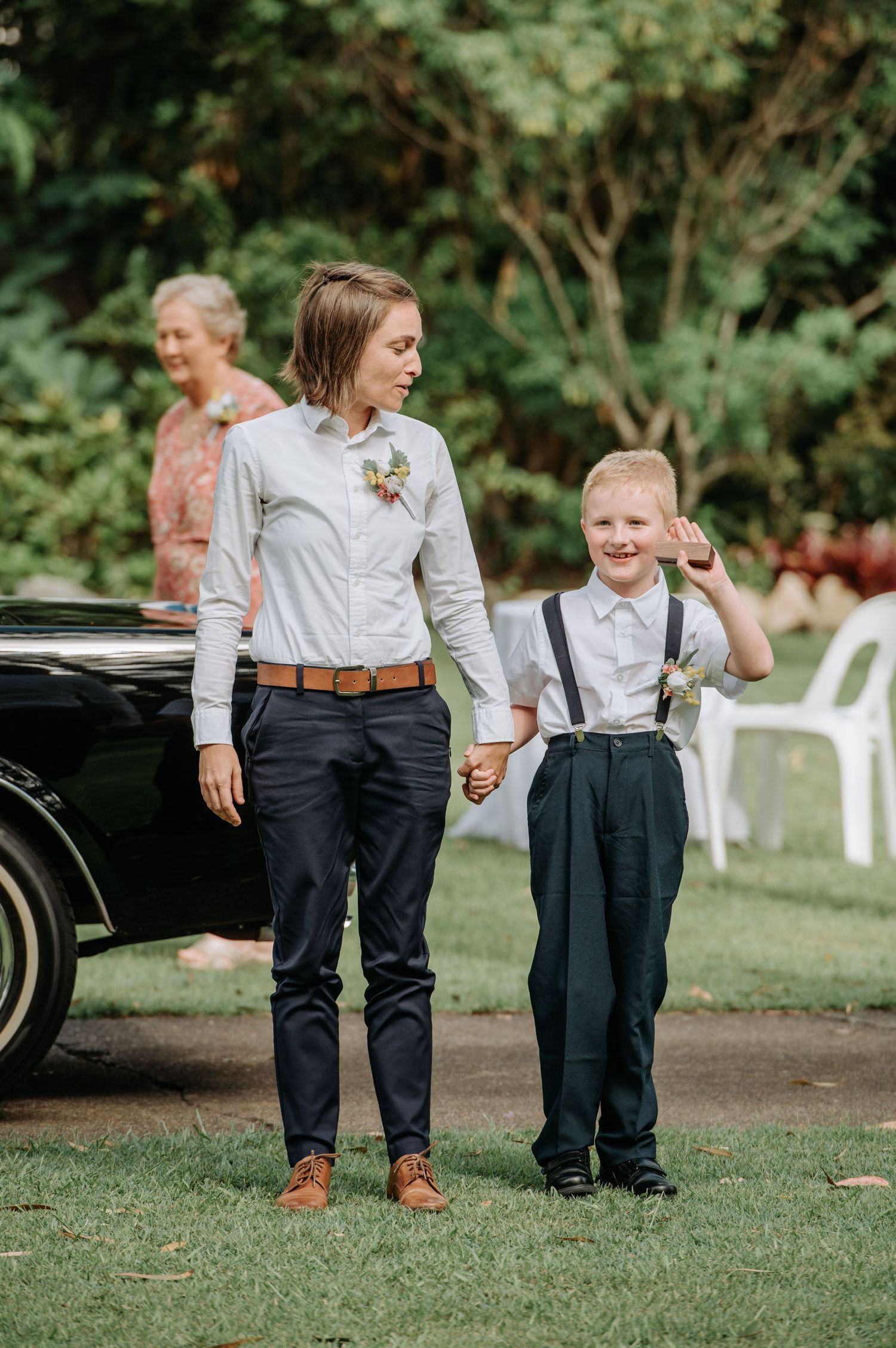 Two people in formal attire standing together on grass near a classic car.