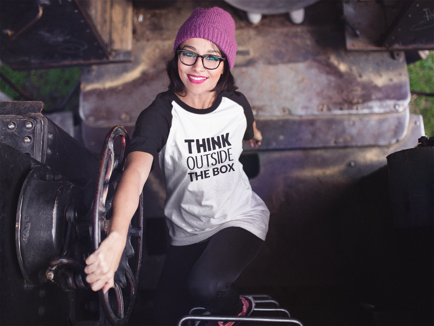 Person in winter beanie and glasses wearing a casual t-shirt in an industrial setting.