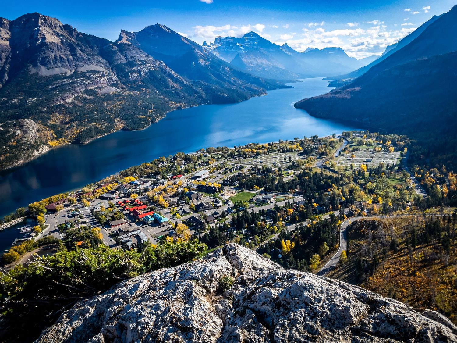 Aerial view of a mountain town nestled along a winding blue lake surrounded by rugged mountain peaks.