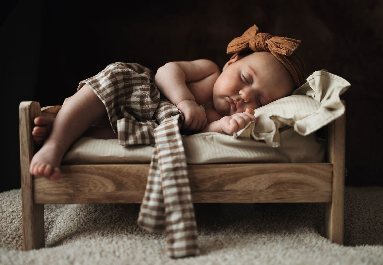 Sleeping newborn baby on a tiny wooden bed with plaid blanket and bow headband in warm tones.
