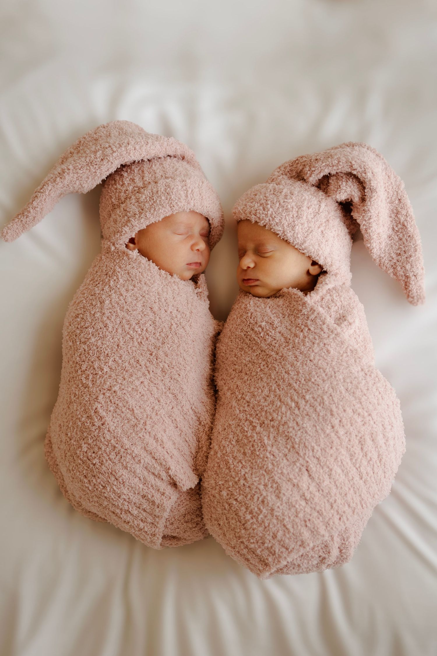 Twin newborns swaddled in matching pink bunny ear wraps sleeping side by side on white background.