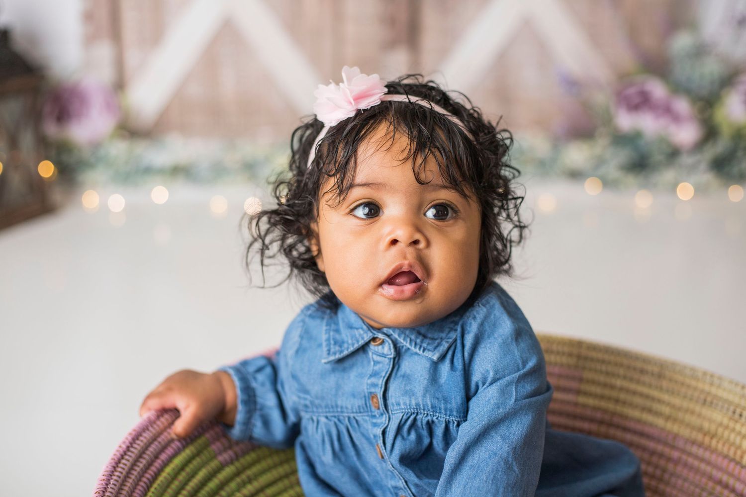 Baby in denim dress sits in woven basket against floral backdrop with vintage lanterns.