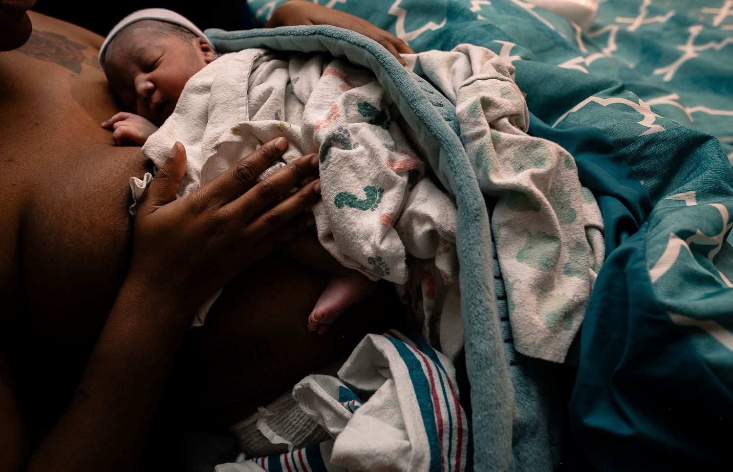 Baby wearing a hat covered with a blanket, lying skin to skin on mother's chest with her hand cradling the baby