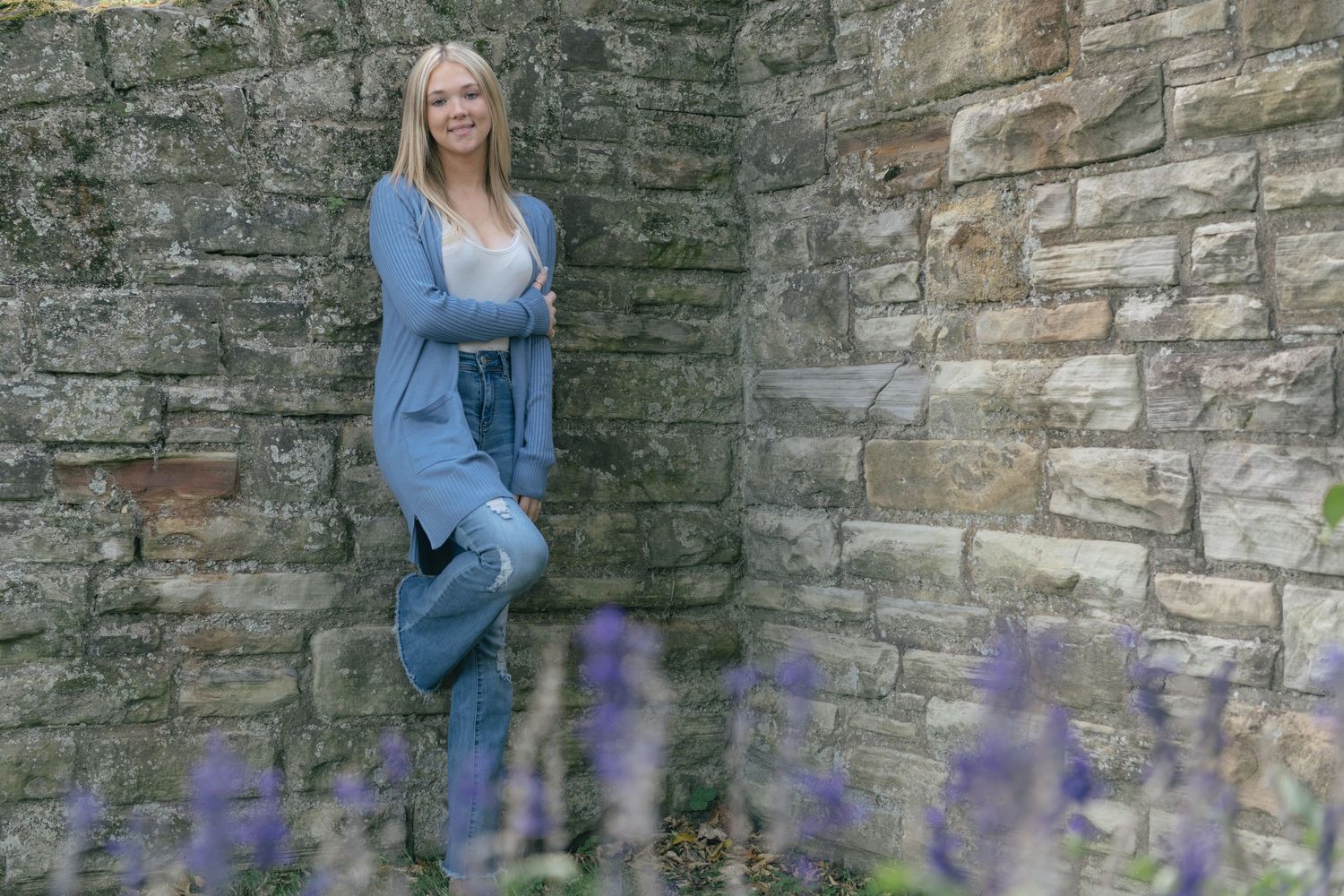Person in blue outfit leaning against stone wall with purple flowers in foreground.