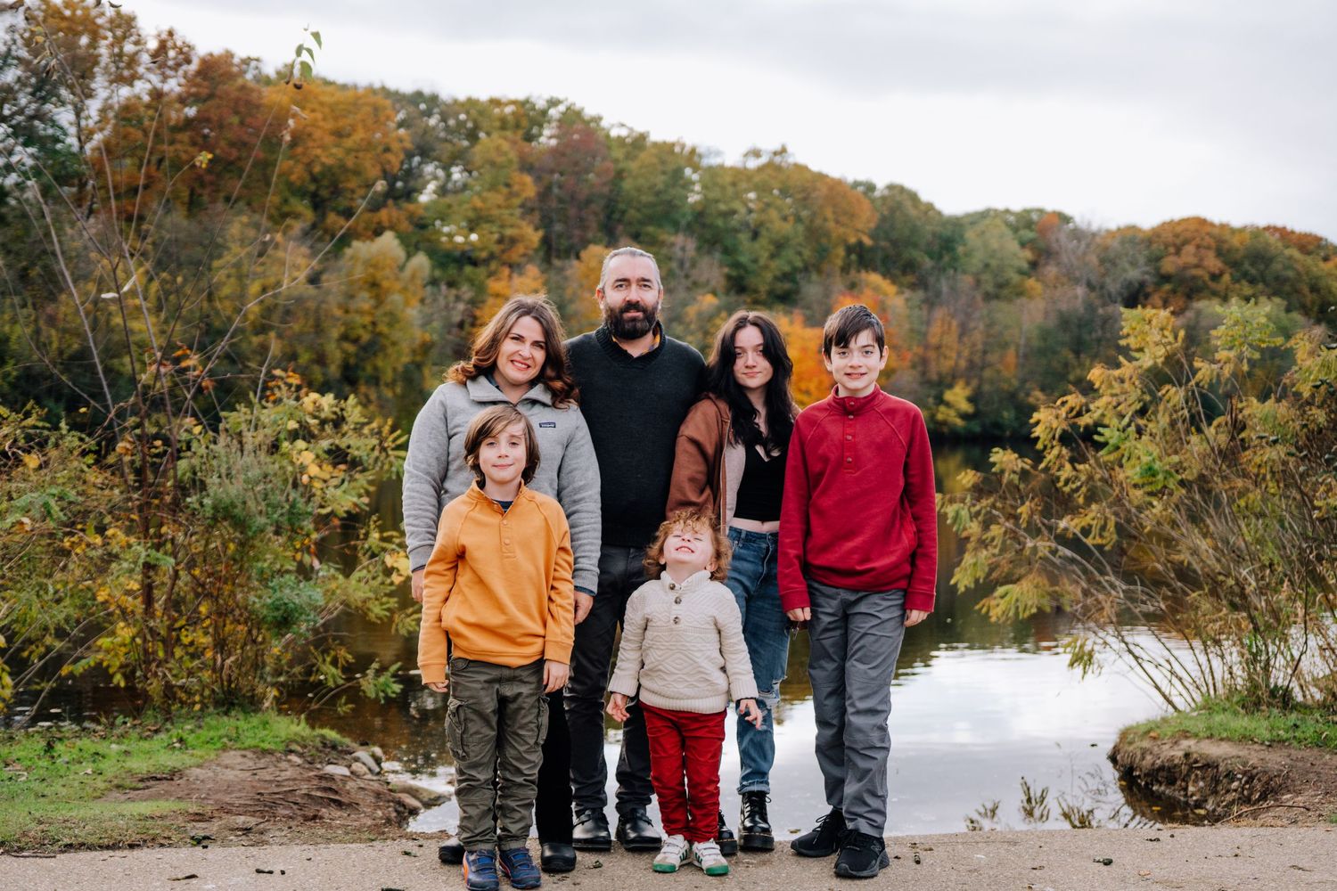 A family group poses together on an autumn day by a lake with fall foliage in the background.