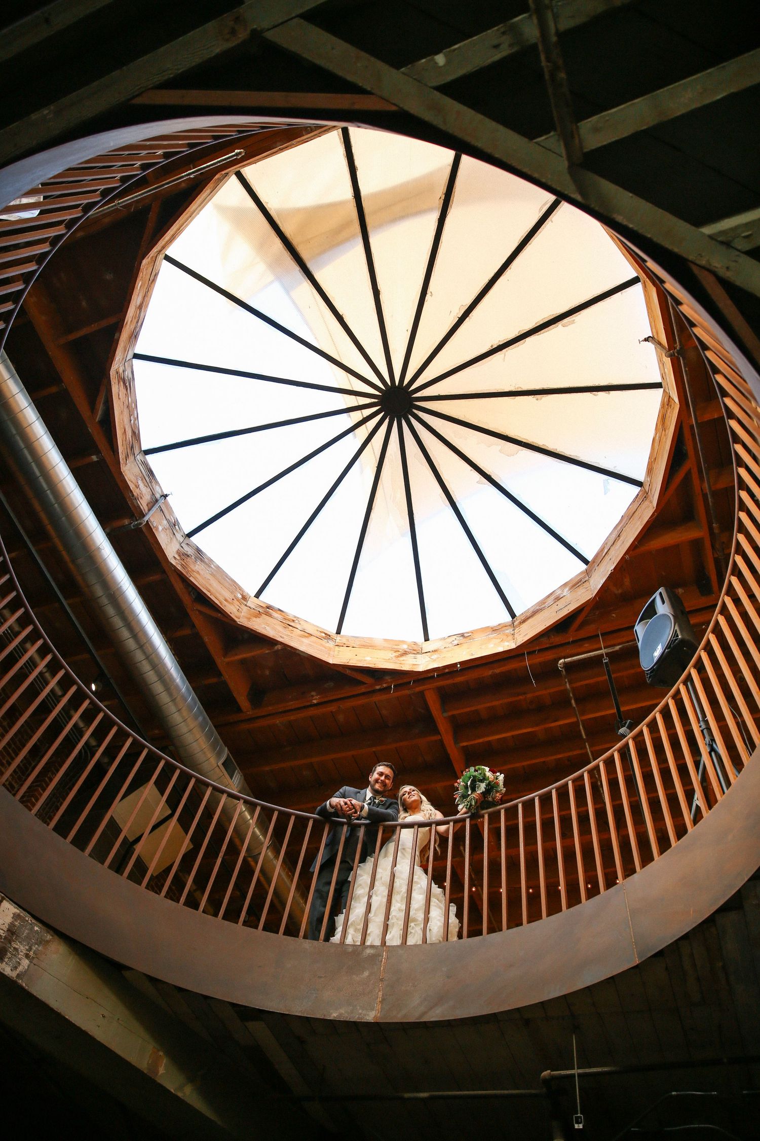 Looking up at a striking circular skylight with wooden beams radiating from center, viewed from curved wooden staircase below.