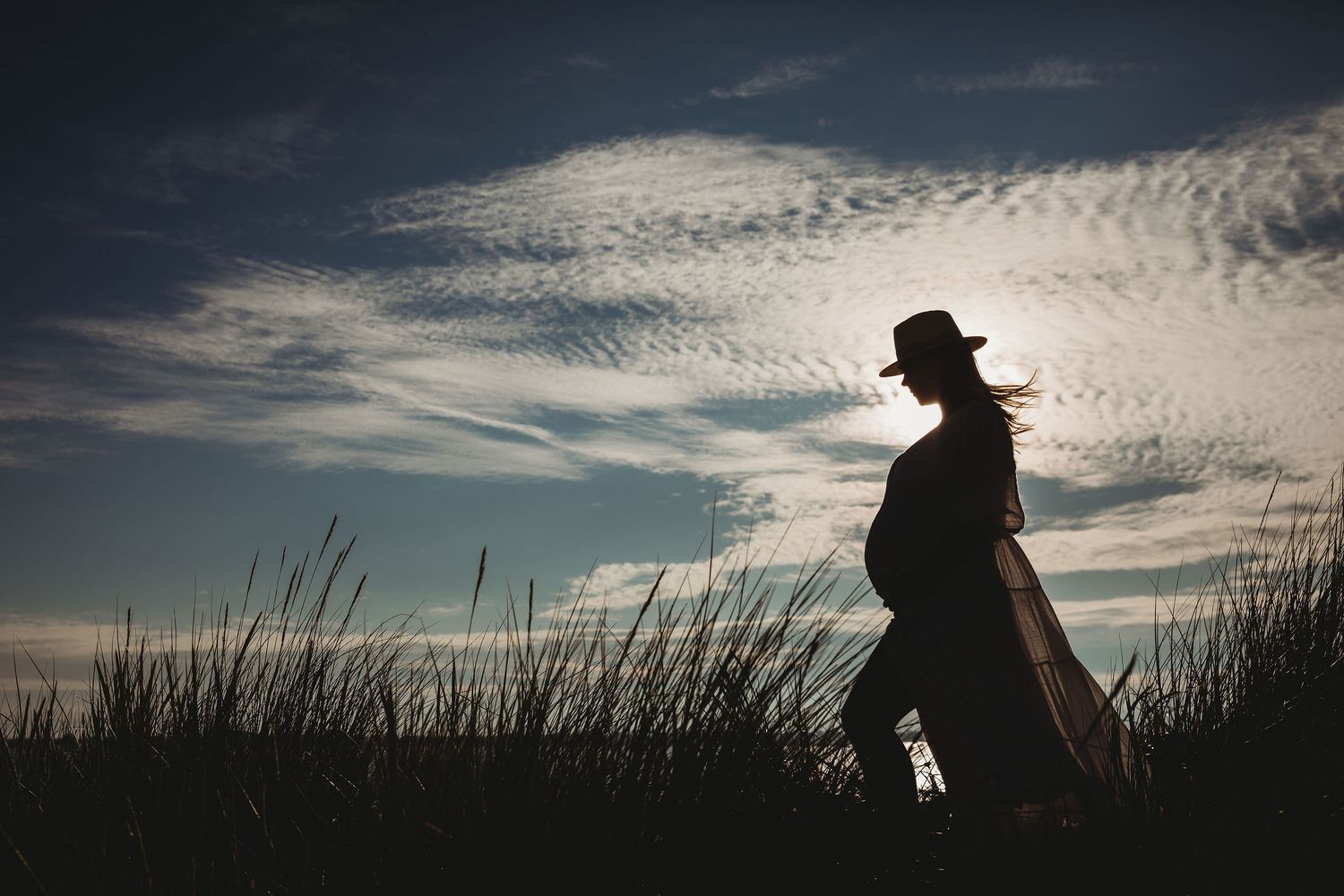 Silhouette of a person in a hat standing among tall grass against a dramatic cloudy sky at sunset.