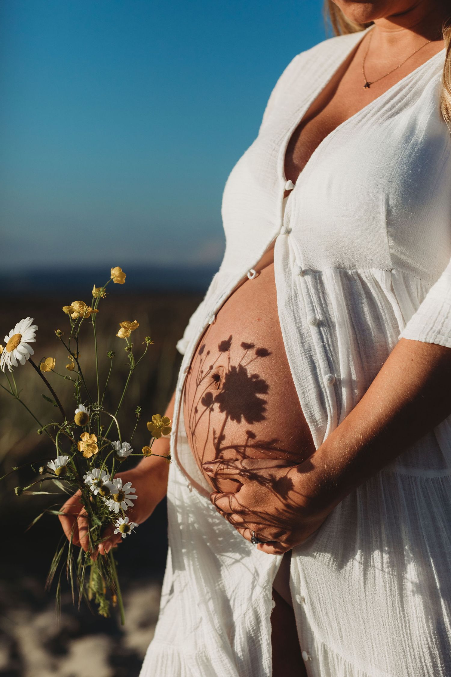Pregnant figure in white dress holding wildflowers casting shadows on belly in golden sunlight.