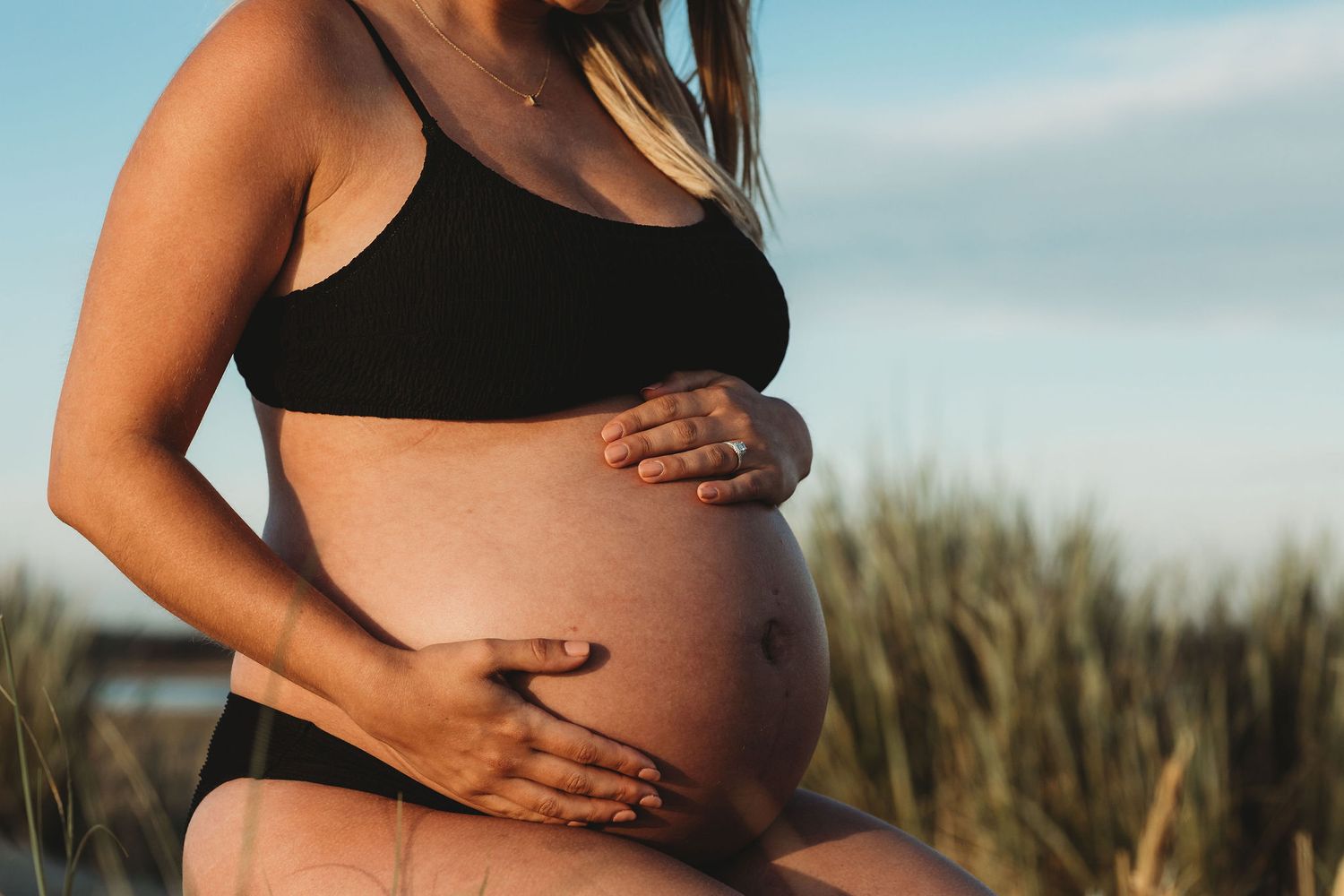 Close-up of pregnant belly with hands resting gently on bare skin against a natural outdoor background.