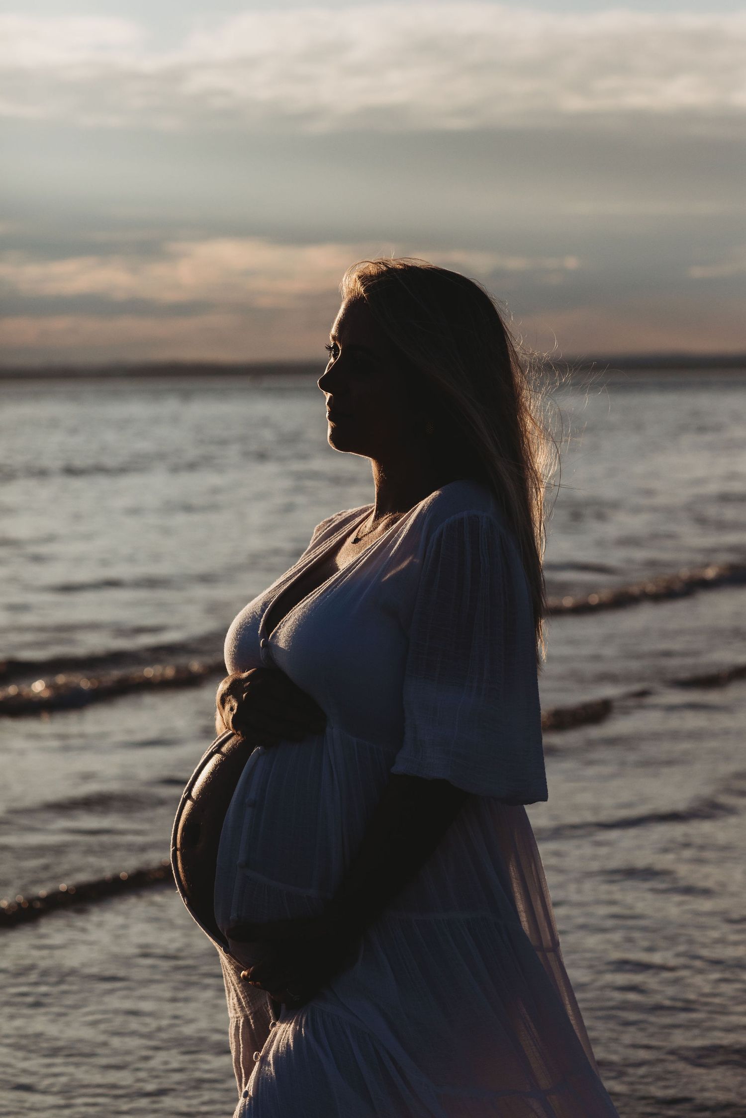 Silhouette of pregnant figure standing by the ocean at sunset in a flowing dress.