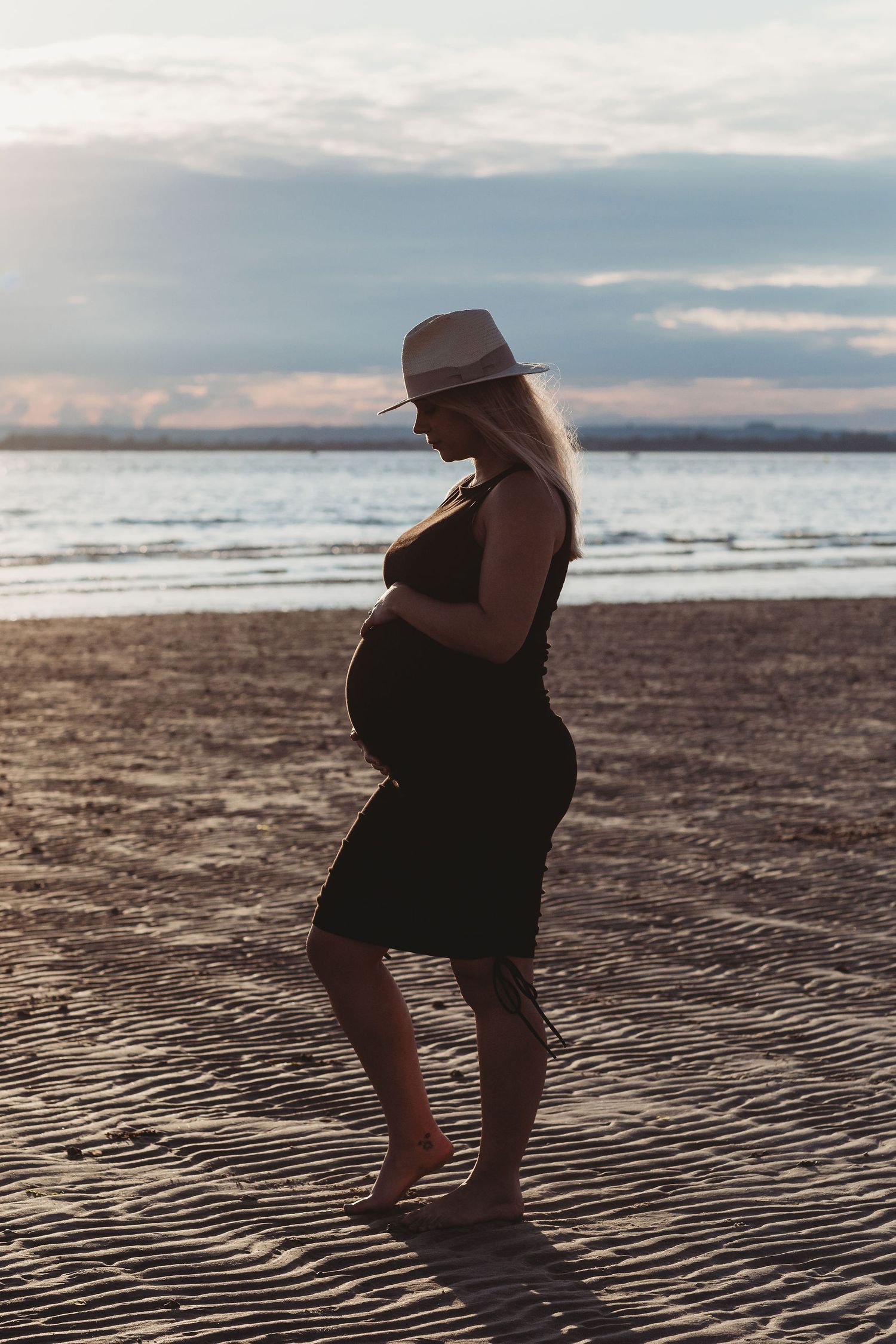 Silhouette of pregnant figure in hat standing on beach at sunset.