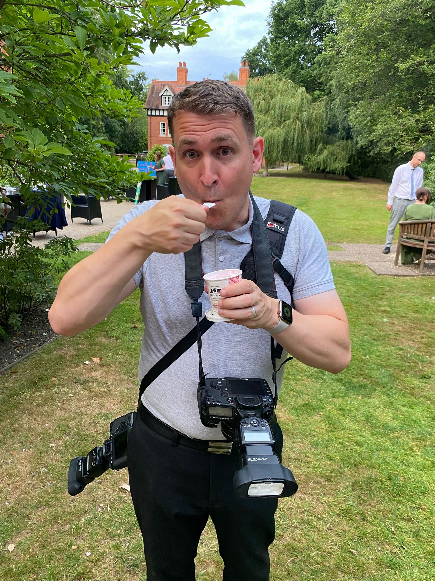 Photographer in gray shirt and black overalls taking pictures in outdoor garden setting.