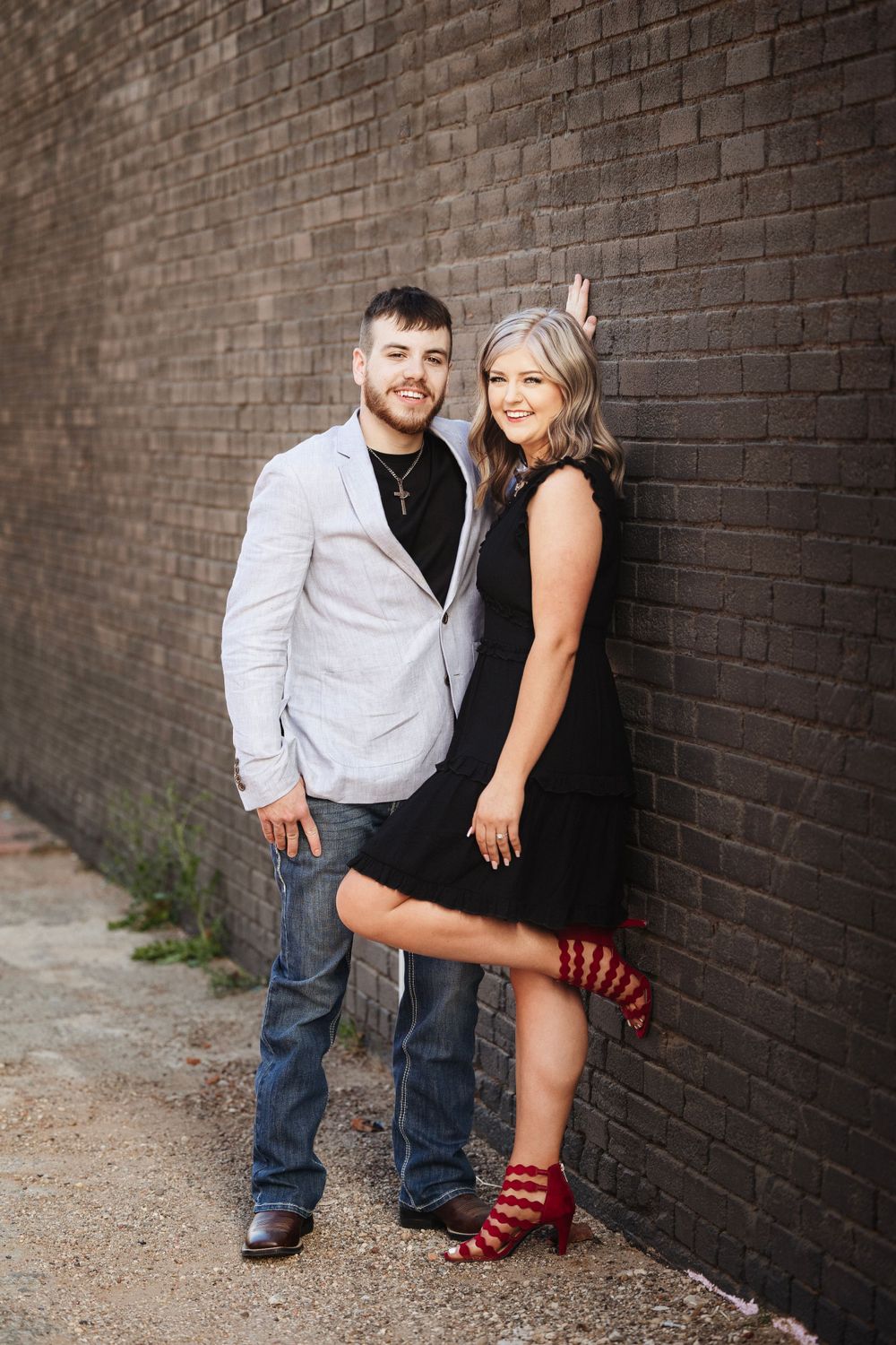 A smiling couple posing against a dark brick wall, man in grey blazer and woman in black dress with red heels.