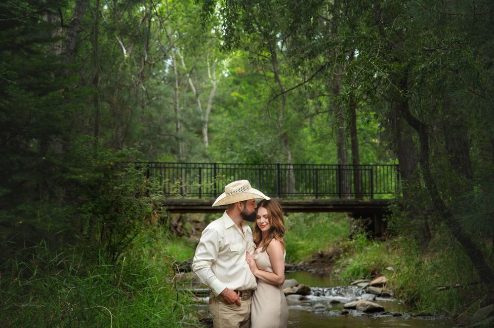 Couple embraces romantically near a creek with a bridge in a lush green forest.
