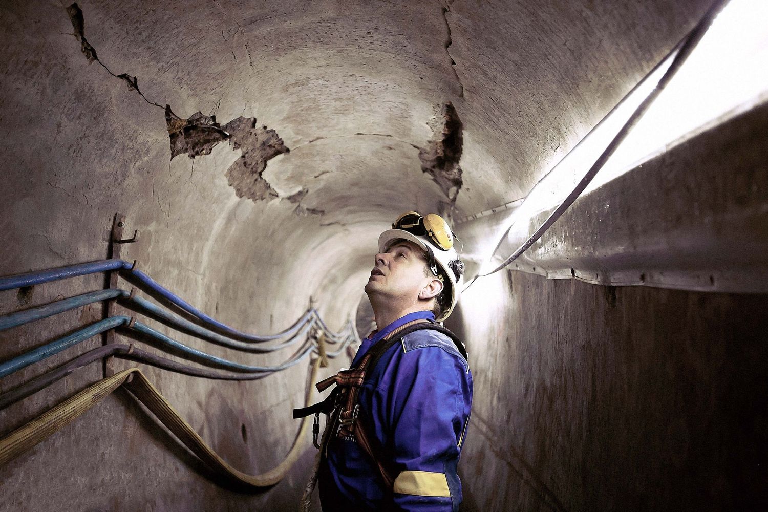 A worker inspects damage in an underground tunnel while wearing safety equipment.