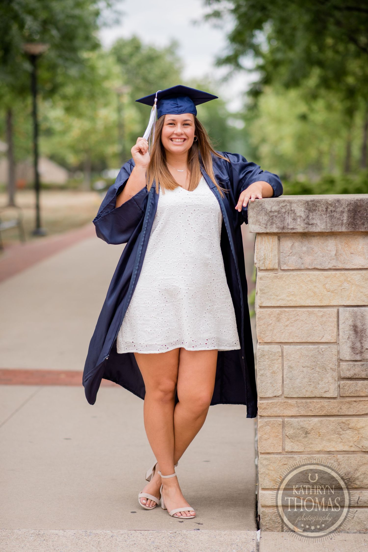 A graduate in blue academic regalia and white dress poses by a stone wall with a bright smile.
