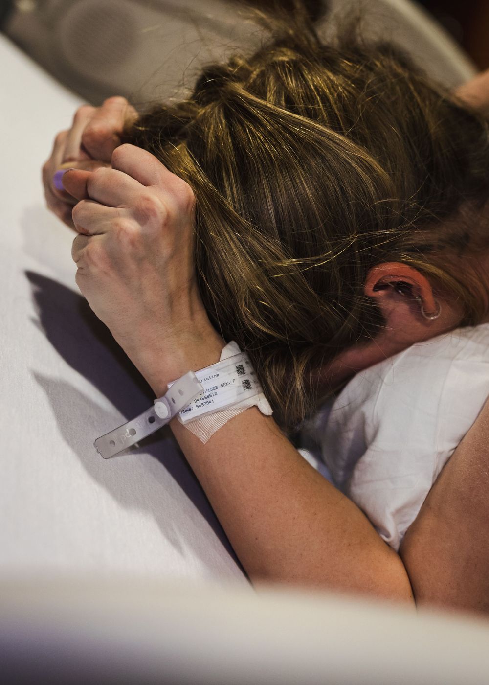 A hospital patient rests with their head down, showing a medical ID bracelet on their wrist.