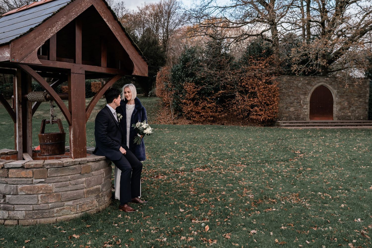 Couple posing by rustic wooden well structure in garden setting.