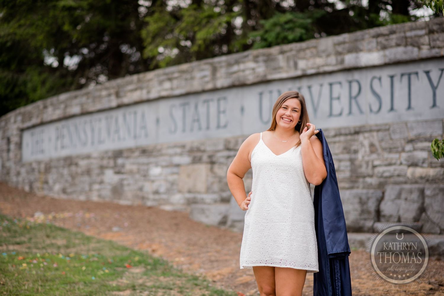 A graduate stands in front of Penn State University's stone sign wearing a white dress.