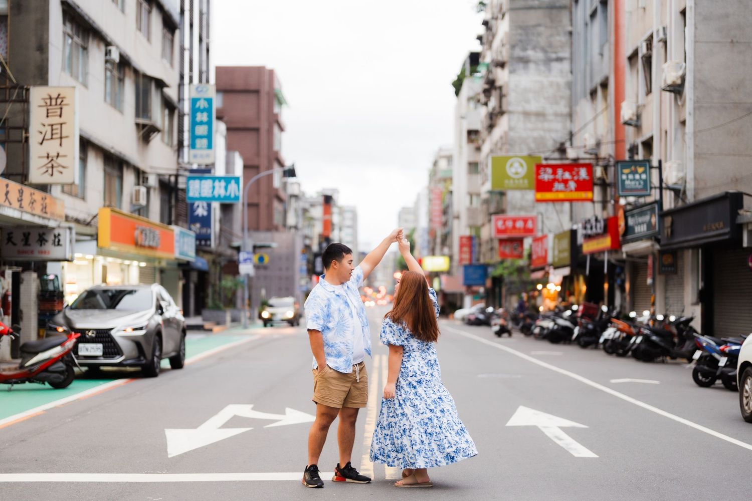 Couple dancing playfully on city street with storefronts and motorcycles in background.