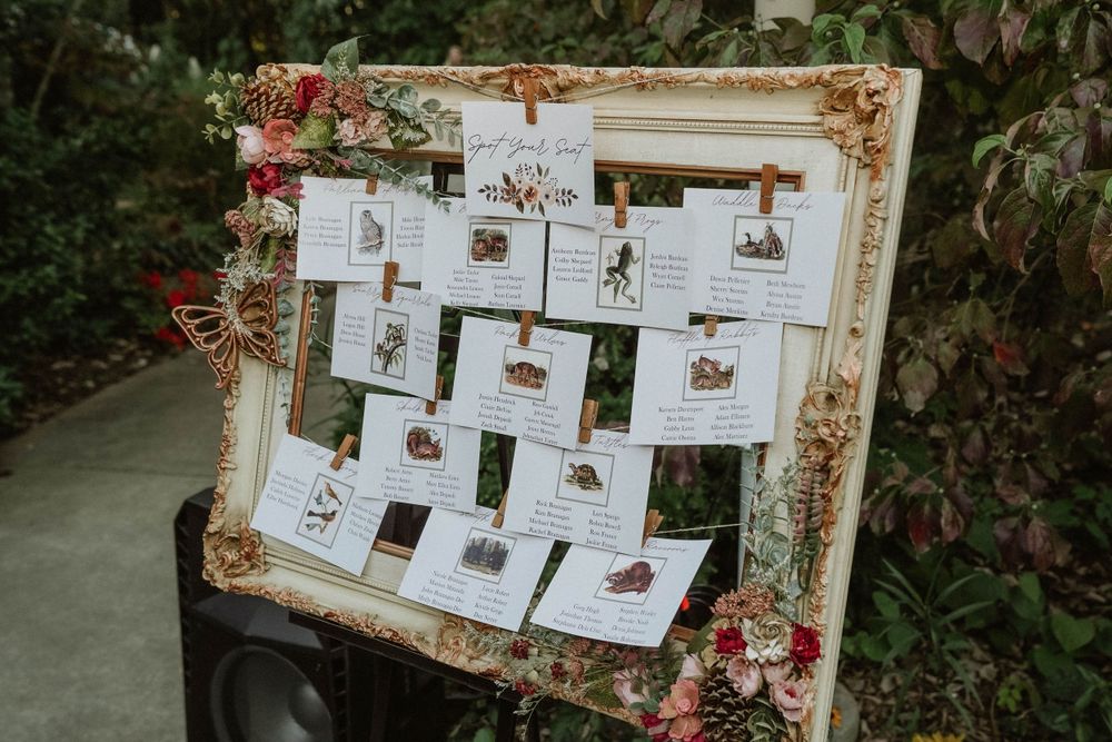 Ornate gold frame decorated with flowers displays wedding seating arrangements on cards.