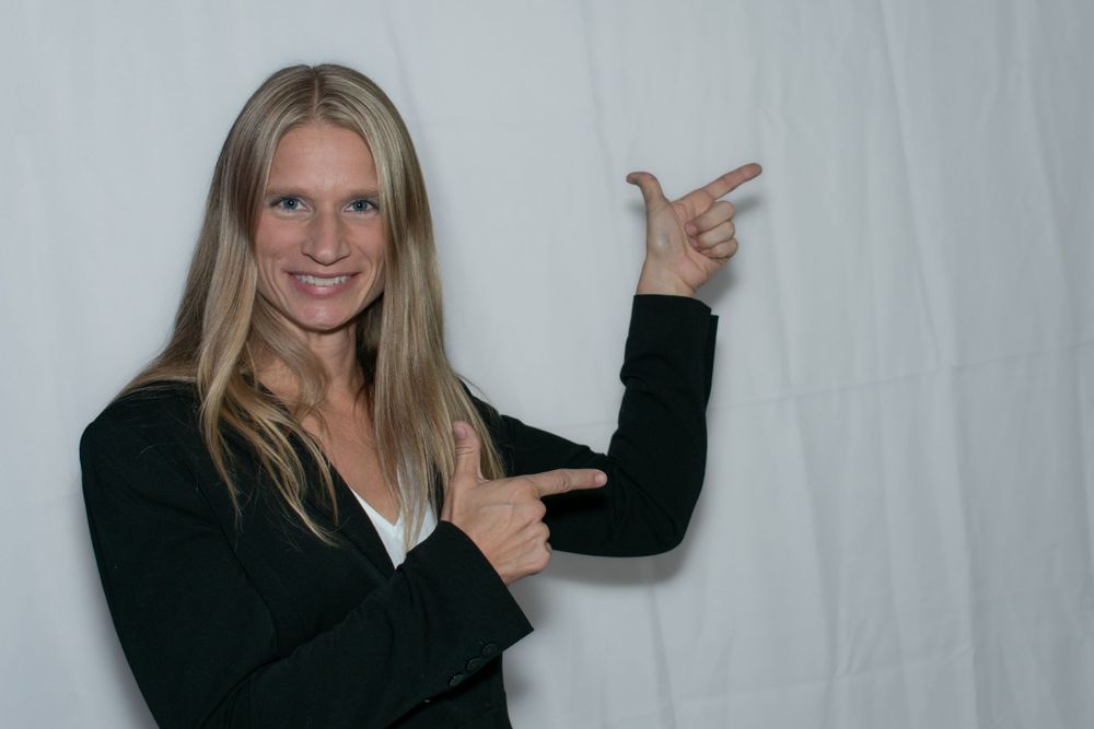 A person in a black blazer points to the side against a white backdrop.