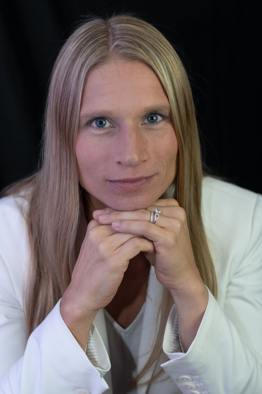 A person in white poses with hands clasped under their chin in a formal portrait.
