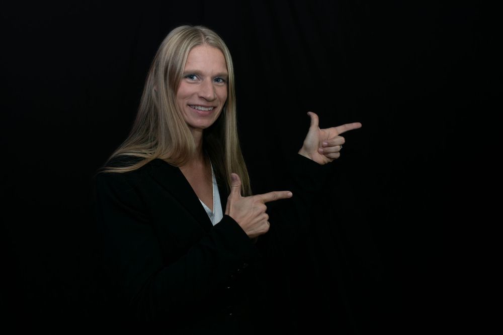 Person in black top pointing with both hands against dark background.