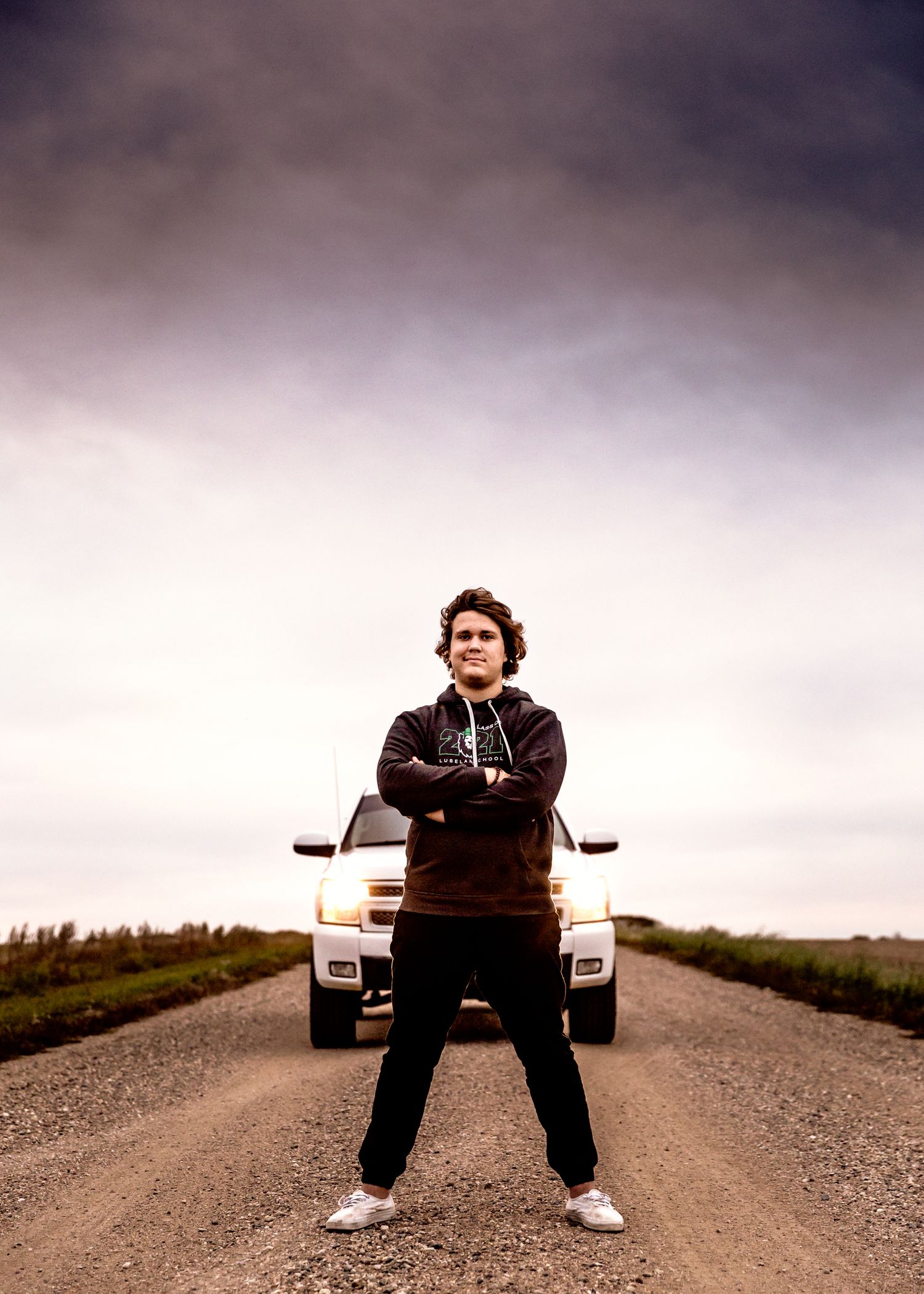 Confident teen standing arms crossed on rural dirt road in front of truck under dramatic sky. Senior grad photos. Graduation photos.