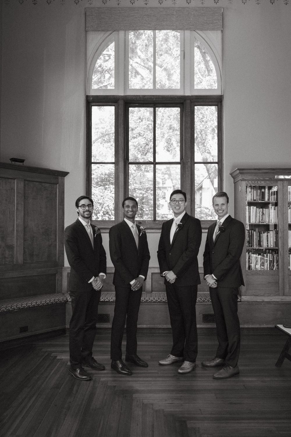 Black and white portrait of groomsmen standing in formal library setting with arched windows.