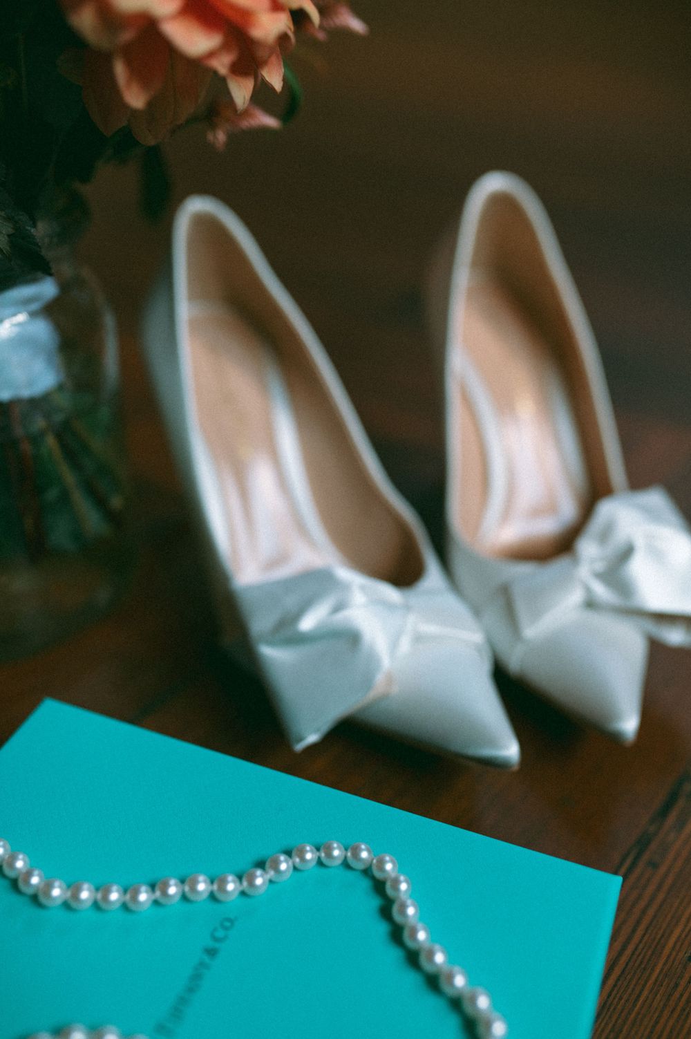 Elegant white bridal shoes with bows displayed next to pearls and a turquoise surface.