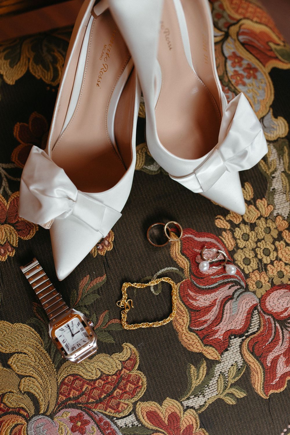 Elegant white bow heels with wedding rings and watch on ornate floral fabric.