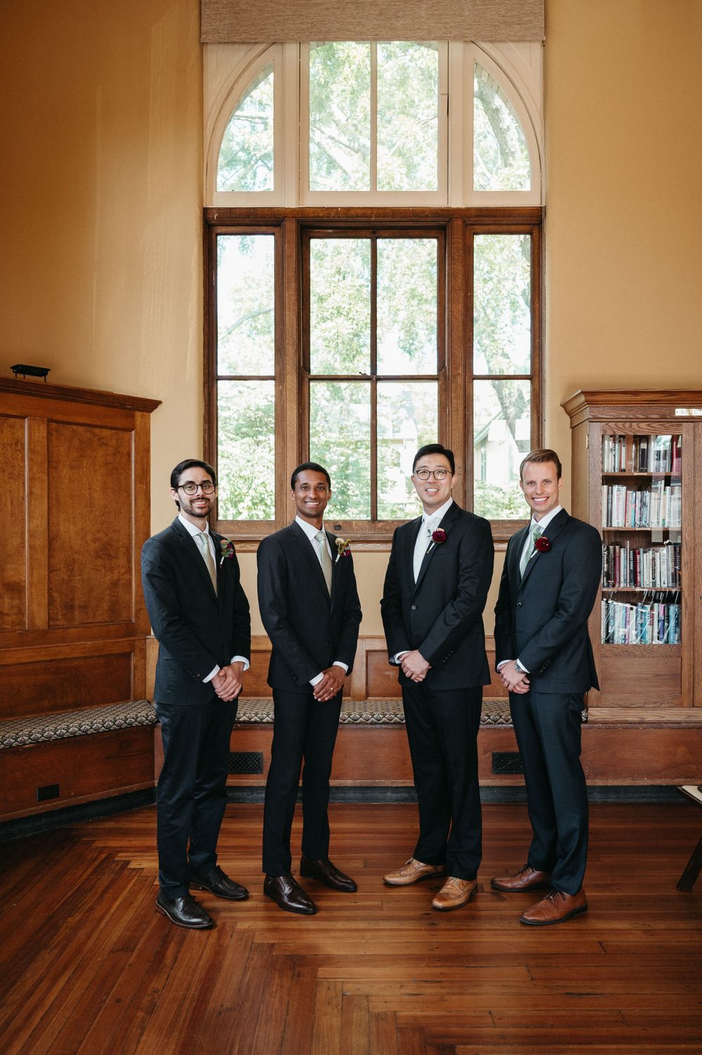 Groomsmen in dark suits pose formally in library with wood paneling and arched windows.
