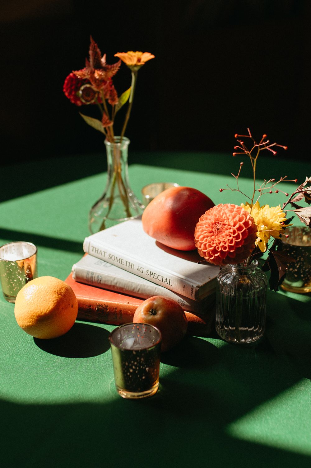 Still life of flowers, citrus fruits and books on green tablecloth with dramatic shadows.
