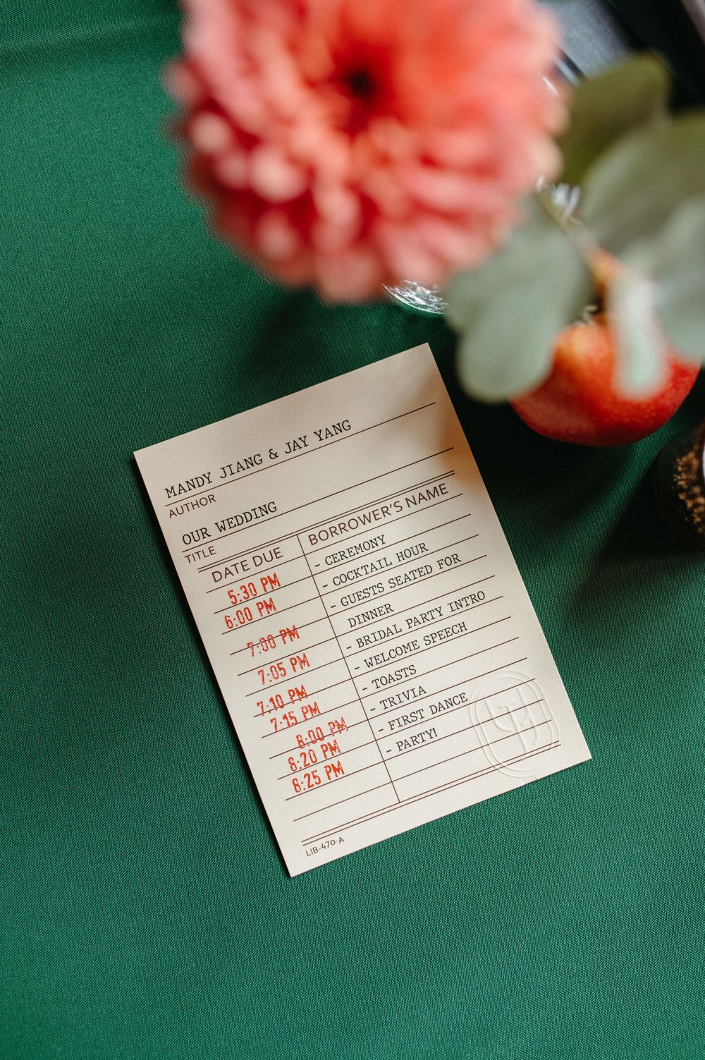Wedding timeline card displayed on green table with coral flower in background.