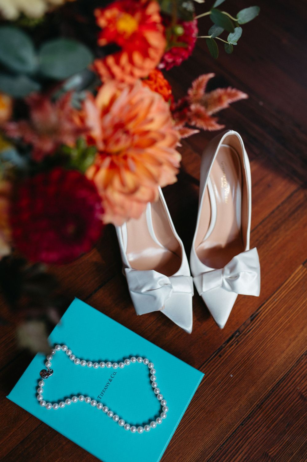 White bow-adorned wedding heels with pearl necklace and coral flowers on wooden surface.