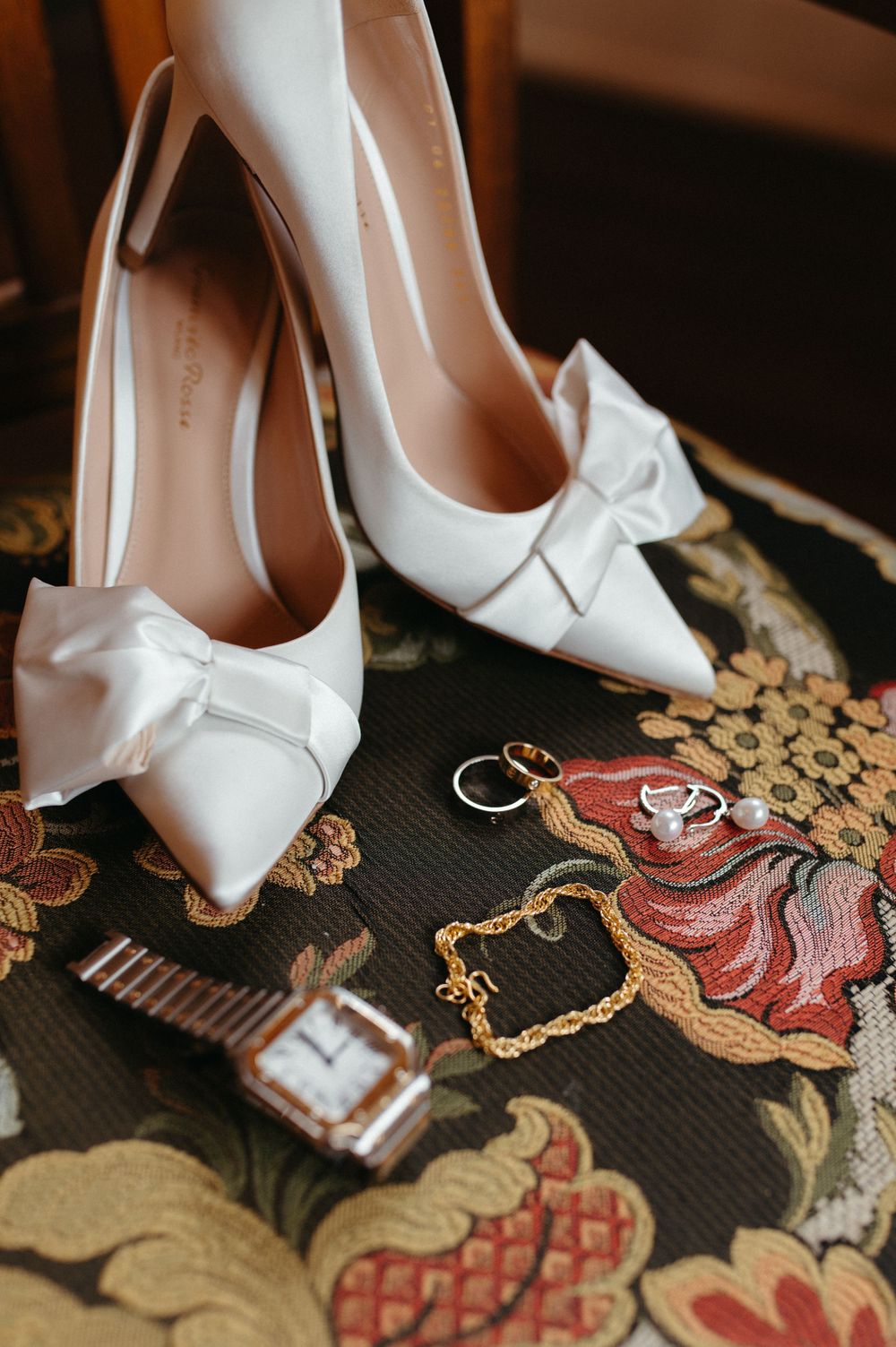 Wedding accessories including white heels, rings, and watch on floral patterned fabric.